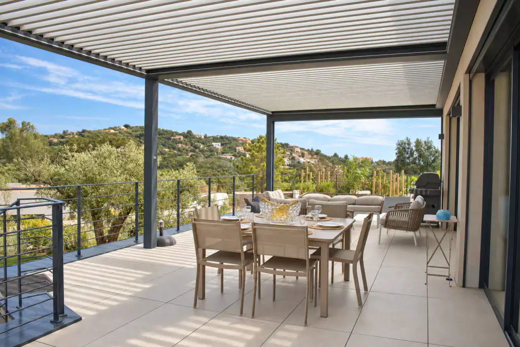 Pergola bioclimatique gris anthracite aux lames entrouvertes couvrant une grande terrasse, table dressée et chaises taupe au centre, garde-corps et collines boisées au fond.