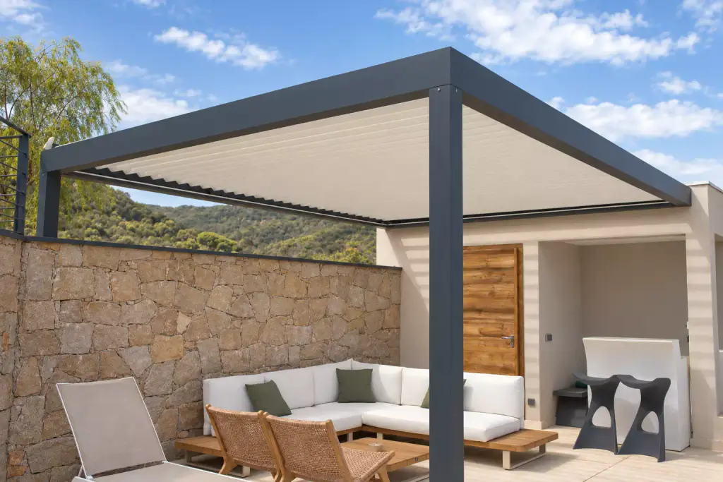 Pergola bioclimatique gris anthracite aux lames claires fermées, vue en contre-plongée, abritant un canapé d’angle blanc et des fauteuils en bois contre un mur de pierre.
