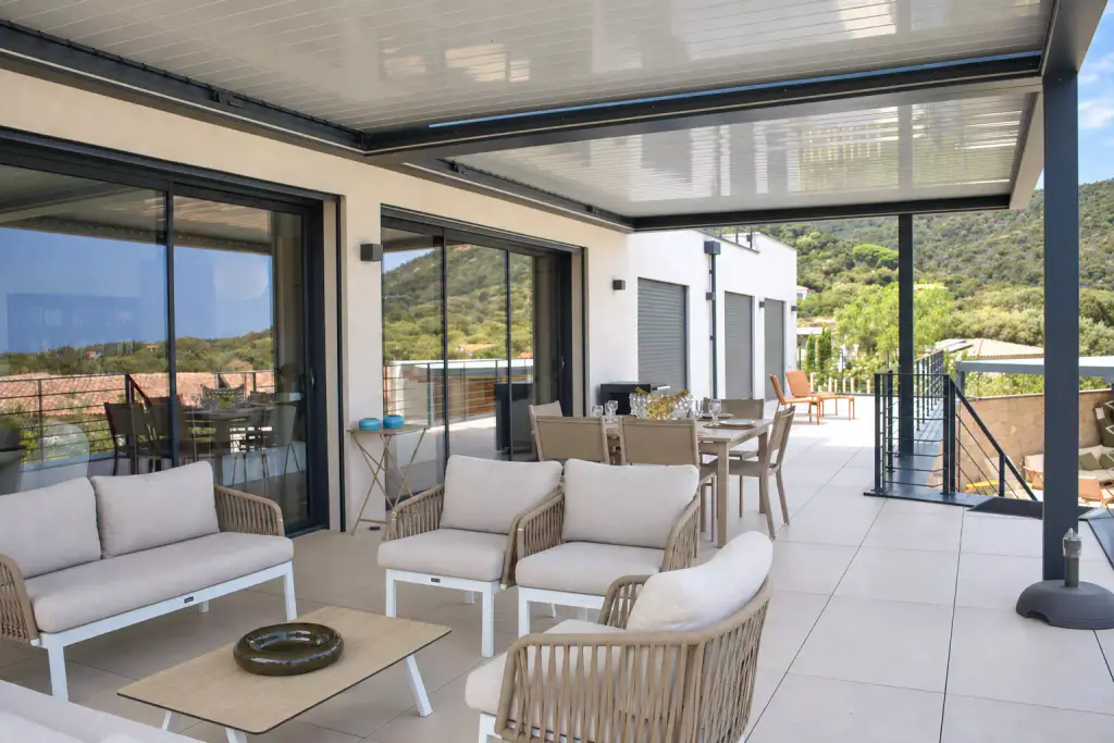 Pergola bioclimatique aux lames fermées couvrant la terrasse : salon en corde tressée et coussins clairs au premier plan, table à manger et baies vitrées coulissantes derrière.