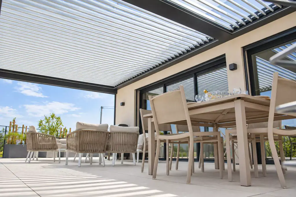 Pergola bioclimatique vue en contre-plongée, lames orientables claires entrouvertes sur le ciel, la table à manger au premier plan.