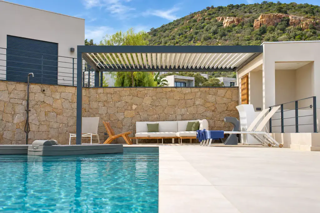 Pergola bioclimatique gris anthracite au bord de la piscine, lames entrouvertes au-dessus d’un canapé d’angle blanc et de bains de soleil, devant un mur de pierre et une colline.