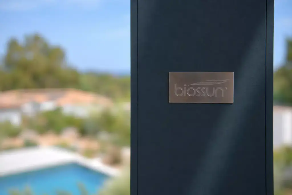 Poteau gris anthracite d’une pergola bioclimatique portant la plaque métallique du fabricant, avec une piscine et des toits de tuiles flous à l’arrière-plan.