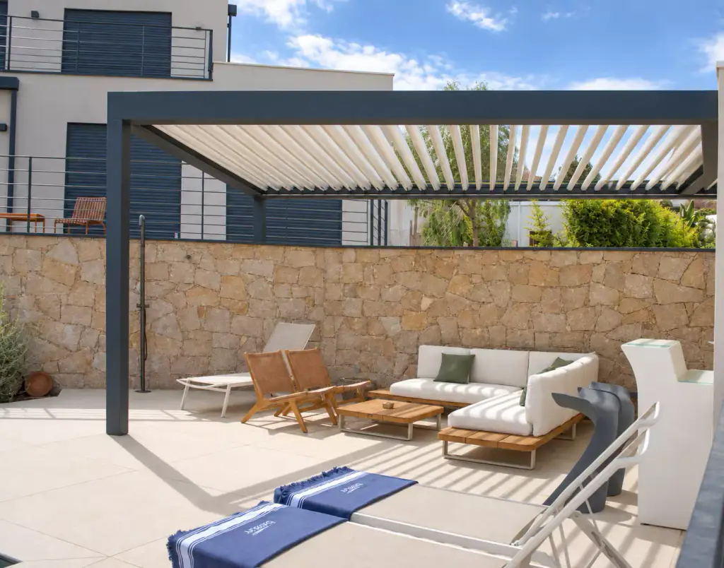 Pergola bioclimatique gris anthracite aux lames ouvertes, face à un mur de pierre, abritant un canapé d’angle blanc et des fauteuils en bois ; bains de soleil au premier plan.