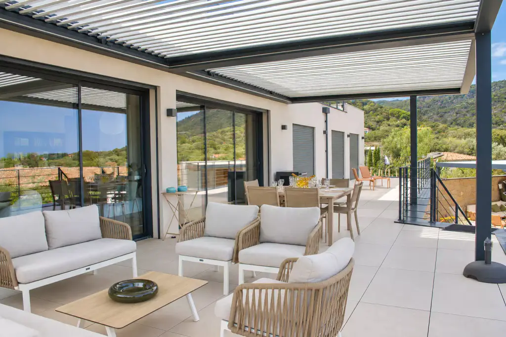 Pergola bioclimatique aux lames entrouvertes couvrant toute la terrasse : salon en corde tressée et coussins clairs au premier plan, table à manger et baies vitrées derrière.