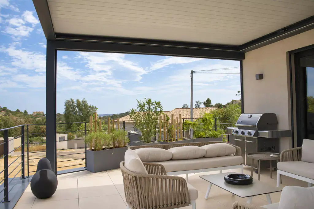 Pergola bioclimatique aux lames fermées, store vertical relevé laissant la vue dégagée, au-dessus d’un salon en corde tressée beige, d’un barbecue à gaz et de jardinières.