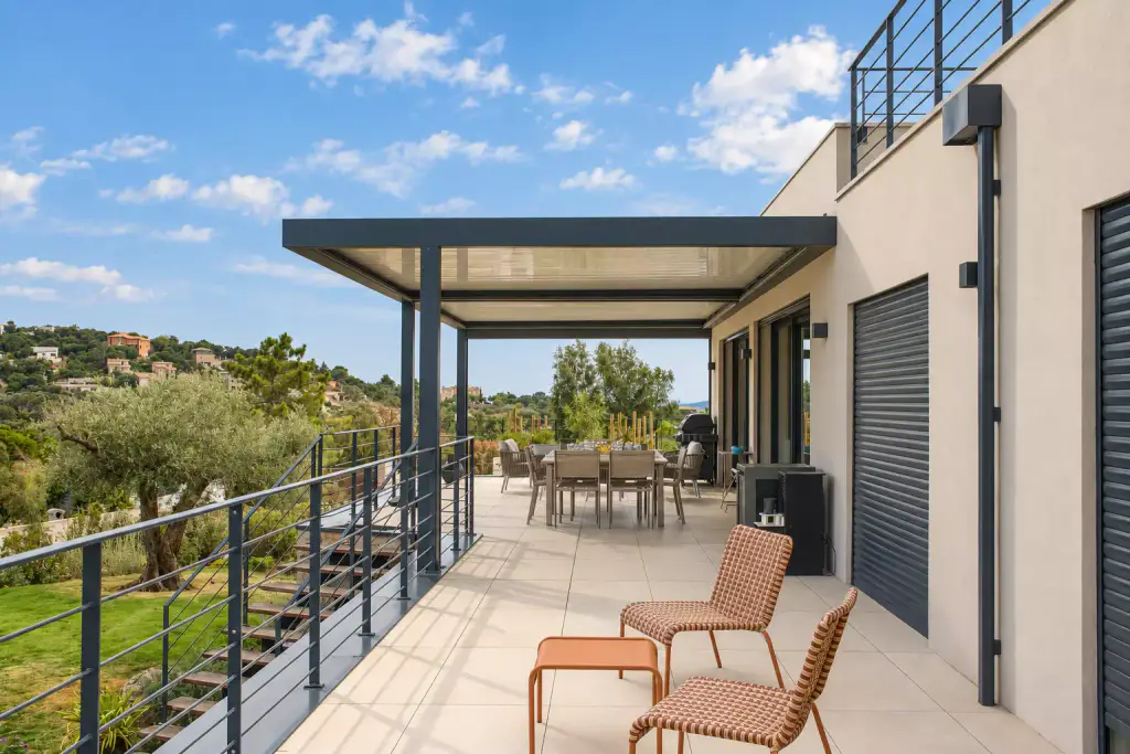 Pergola bioclimatique gris anthracite adossée à une villa contemporaine, au bout d’une terrasse à garde-corps ouverte sur les collines, fauteuils terracotta au premier plan.