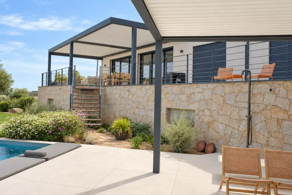 Pergola bioclimatique de la terrasse haute vue depuis celle de la piscine, soubassement de pierre, douche extérieure, fauteuils en bois et jardin de rocaille entre les deux.