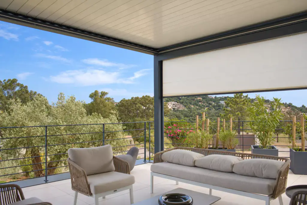 Store screen clair à mi-hauteur sur un côté d’une pergola bioclimatique, salon en corde tressée beige au-dessous, l’autre côté ouvert sur des oliviers et des collines.
