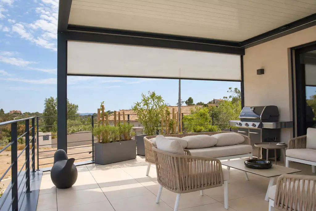 Store screen clair à moitié descendu entre deux poteaux d’une pergola bioclimatique gris anthracite, au-dessus d’un salon en corde tressée et de jardinières plantées.