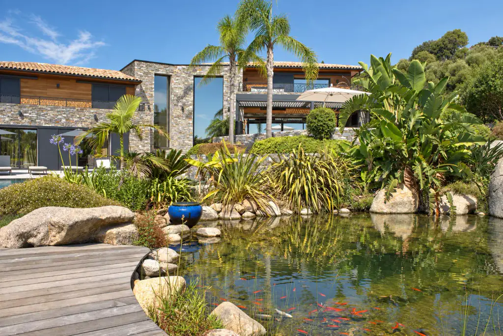 Bassin d’agrément où nagent des poissons rouges, ponton en bois, rochers et plantes luxuriantes, devant une villa en pierre et bois entourée de palmiers.
