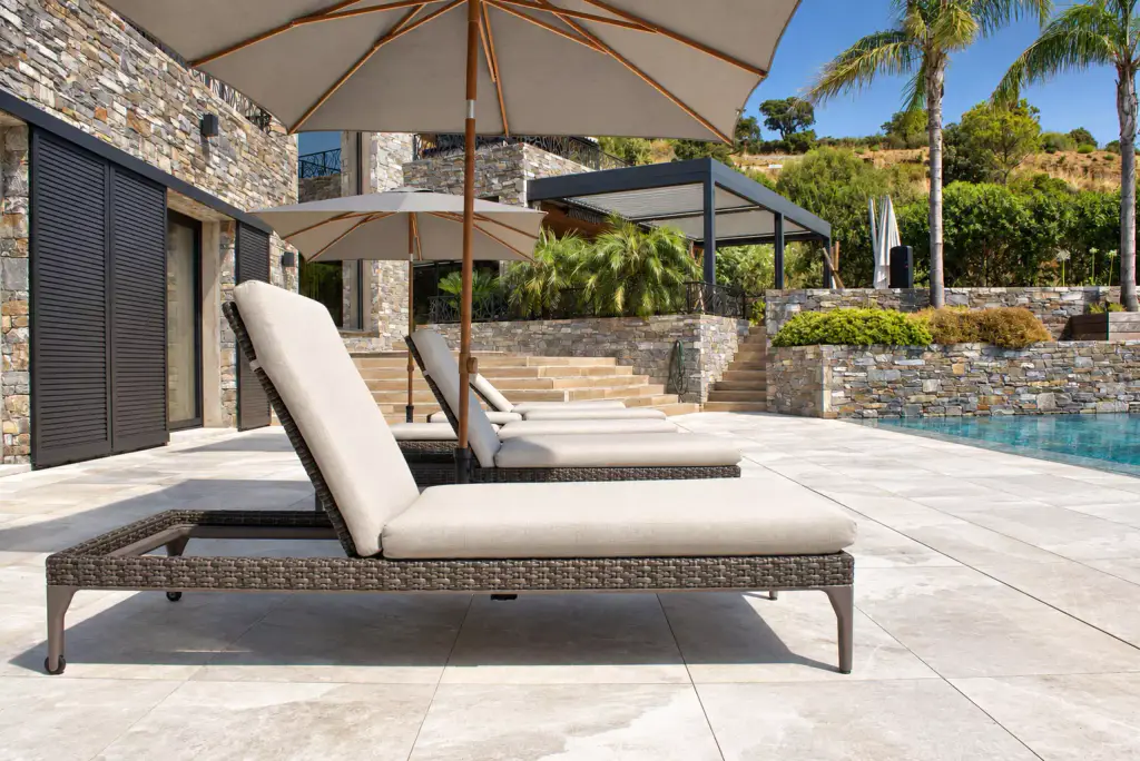 Bain de soleil tressé à matelas beige sous un parasol clair à mât en bois, au bord d’une piscine ; pergola gris anthracite et murets de pierre à l’arrière-plan.