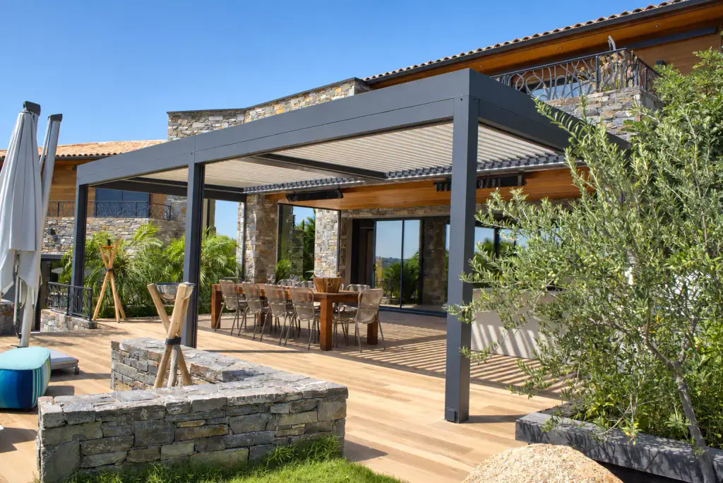 Pergola bioclimatique gris anthracite adossée à une villa en pierre et bois, abritant une longue table et ses chaises en corde, muret de pierre sèche et olivier au premier plan.