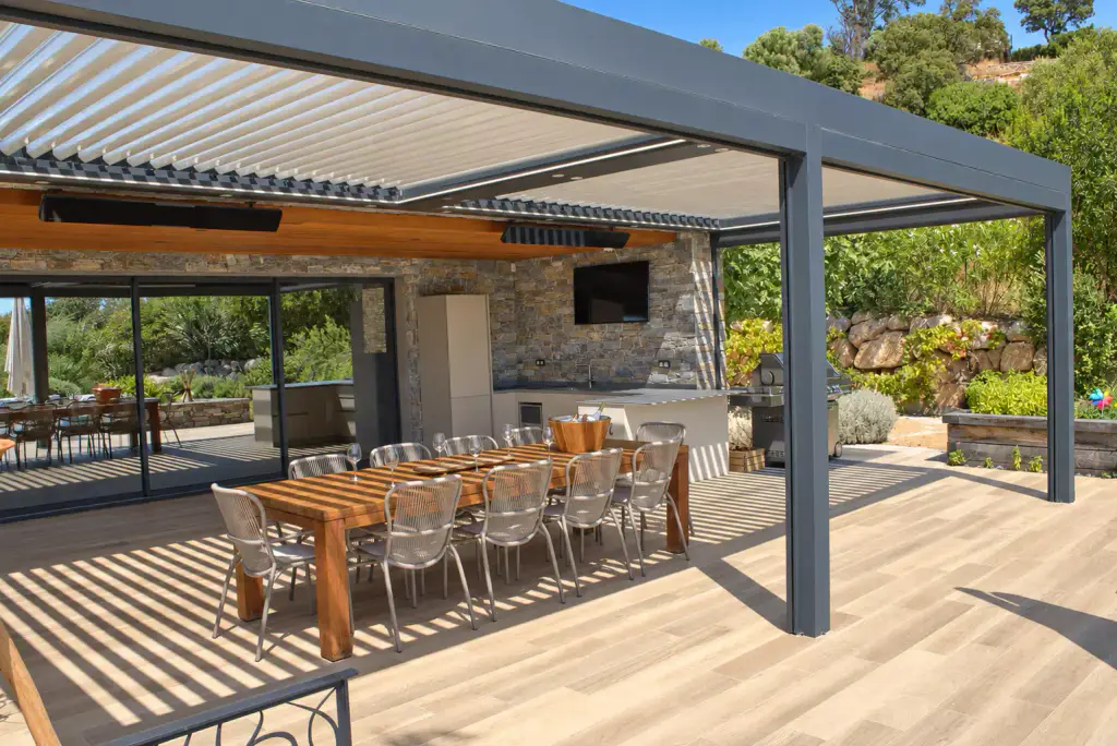 Pergola bioclimatique gris anthracite aux lames entrouvertes couvrant une cuisine d’été en pierre et une longue table en bois, chaises en corde et barbecue en inox.