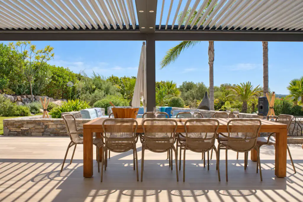 Pergola bioclimatique aux lames ouvertes projetant des ombres rayées sur le sol, grande table en bois massif entourée de chaises en corde tressée, palmiers et jardin au fond.