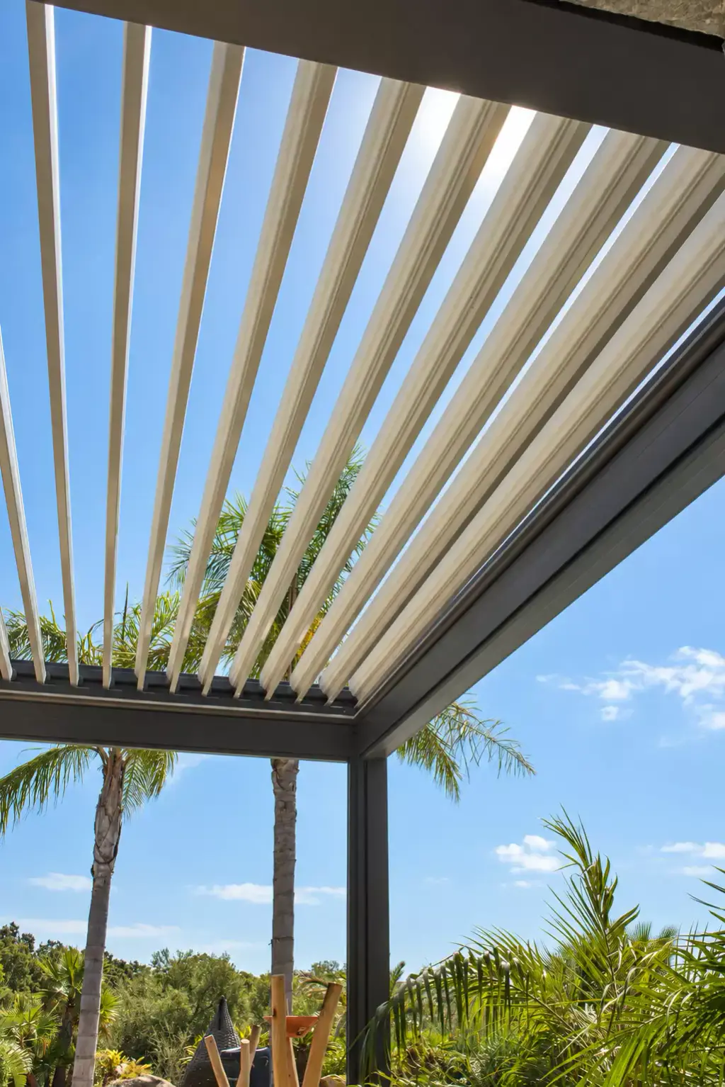 Lames orientables claires d’une pergola bioclimatique grandes ouvertes sur le ciel bleu, vues par-dessous, avec l’angle de la structure gris anthracite et un palmier.