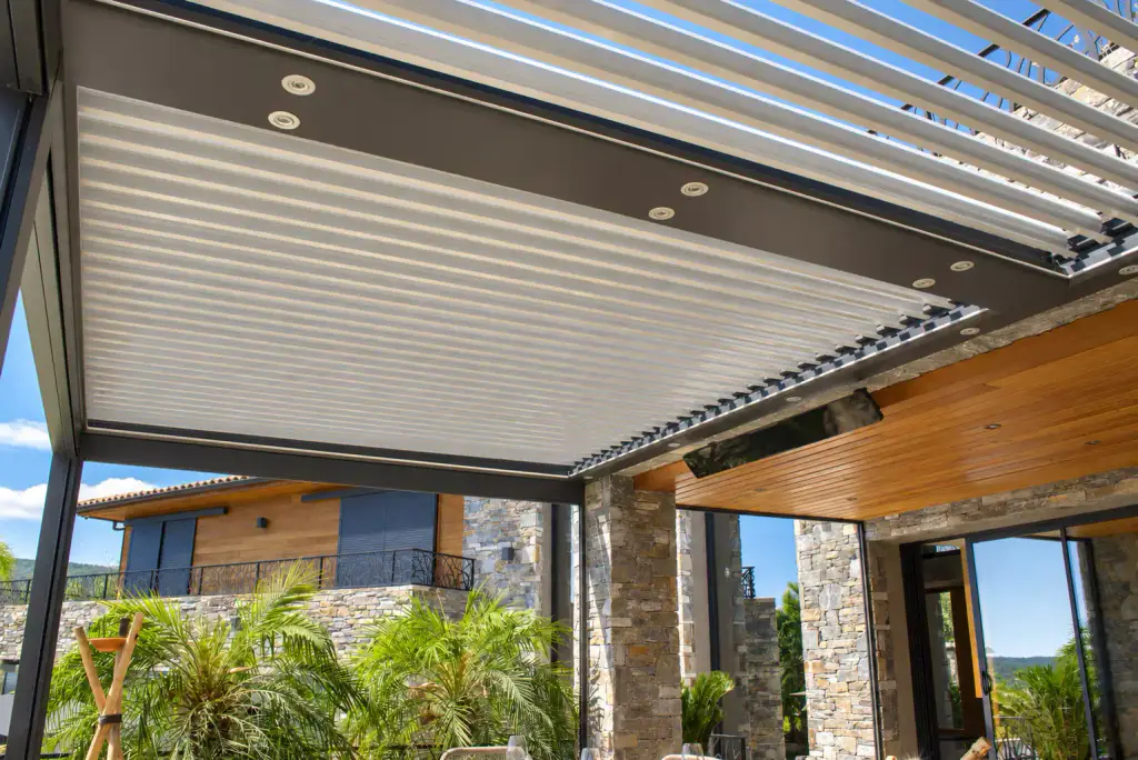 Sous-face d’une pergola bioclimatique : lames claires en partie ouvertes, spots intégrés dans la structure gris anthracite, auvent en bois derrière.