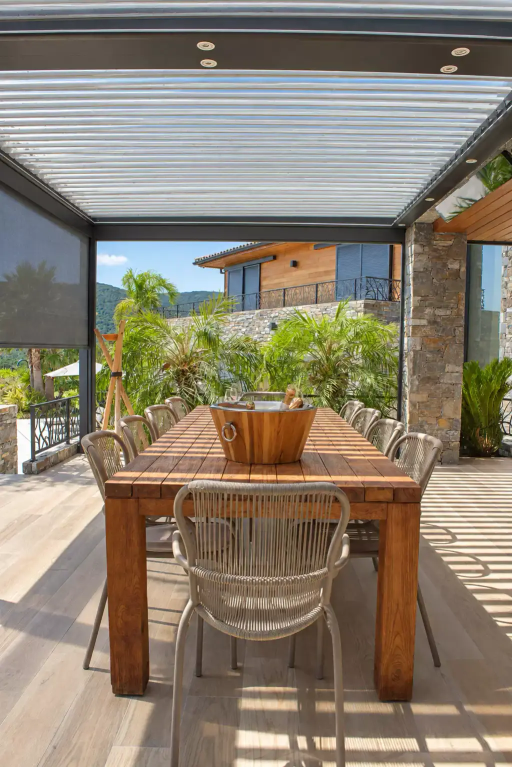 Pergola bioclimatique aux lames entrouvertes et store screen gris descendu sur la gauche, longue table en bois vue en bout, chaises en corde et villa en pierre au fond.