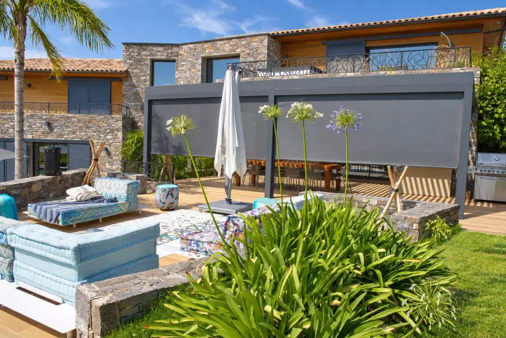 Casa Palma vue du jardin : la pergola gris anthracite, stores screen descendus, devant la villa de pierre et de bois.