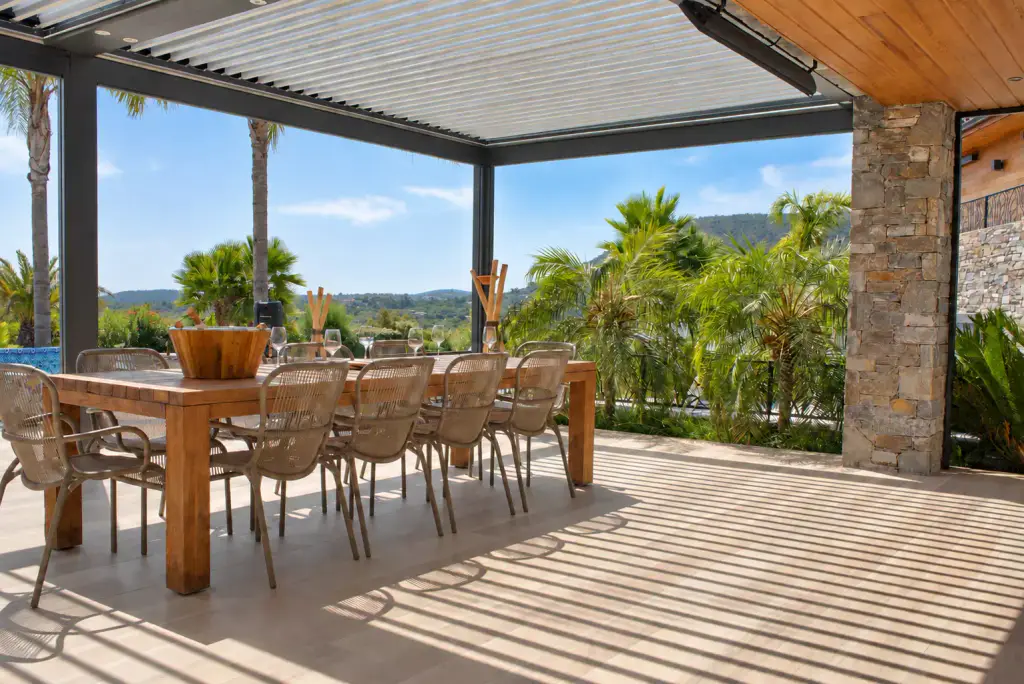 Pergola bioclimatique aux lames entrouvertes au-dessus d’une longue table en bois massif et de chaises en corde tressée, pilier de pierre à droite, palmiers et collines au fond.
