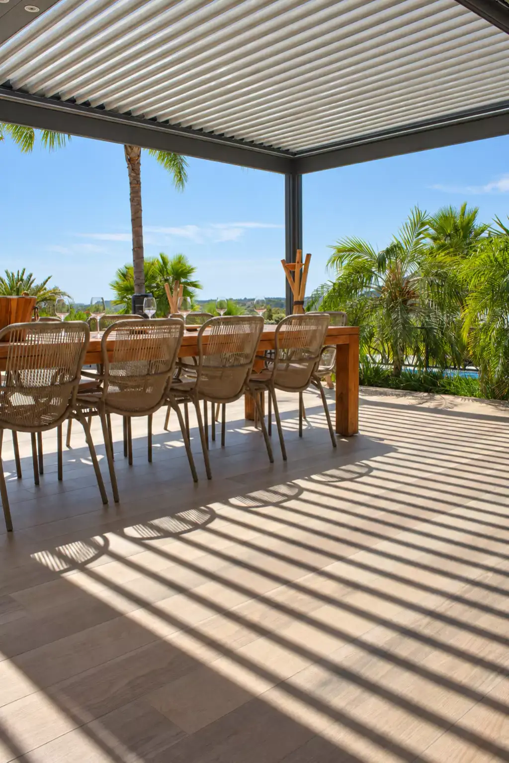 Pergola bioclimatique aux lames ouvertes dessinant des ombres rayées sur le sol aspect bois, longue table en bois et chaises en corde tressée devant des palmiers.