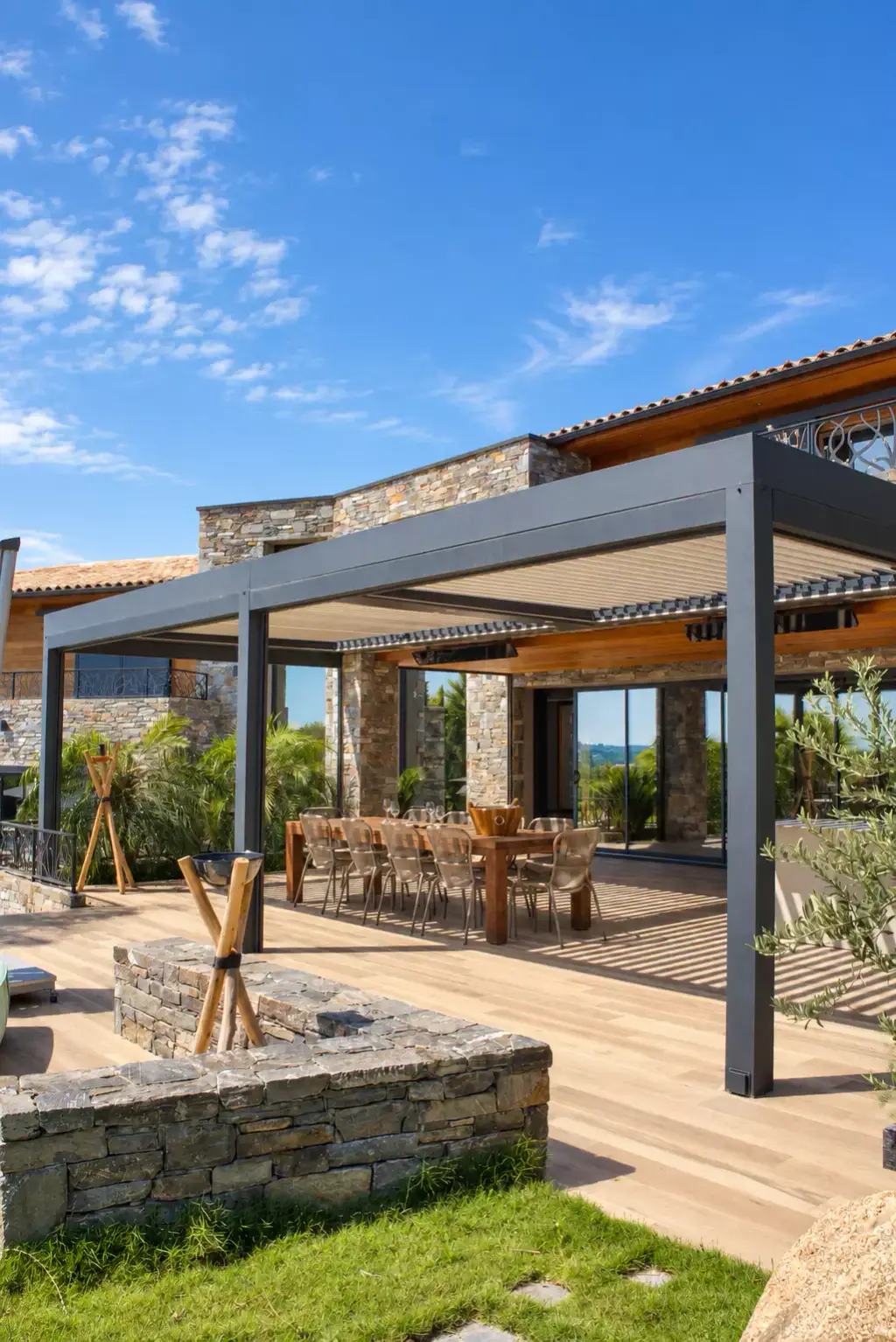 Pergola bioclimatique gris anthracite adossée à une villa en pierre et bois, longue table et chaises en corde dessous, muret de pierre sèche et pelouse au premier plan.