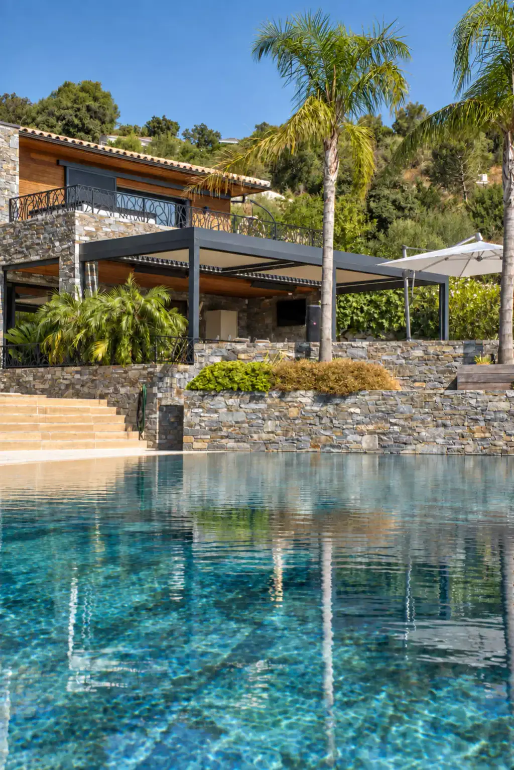 Piscine à l’eau turquoise où se reflètent les palmiers, marches et murets de pierre sèche, et sur la terrasse haute la pergola bioclimatique gris anthracite de la villa.
