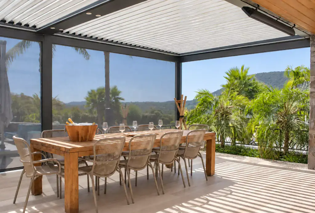Stores screen gris descendus sur un côté d’une pergola bioclimatique, laissant deviner le paysage, au-dessus d’une longue table en bois et de chaises en corde tressée.