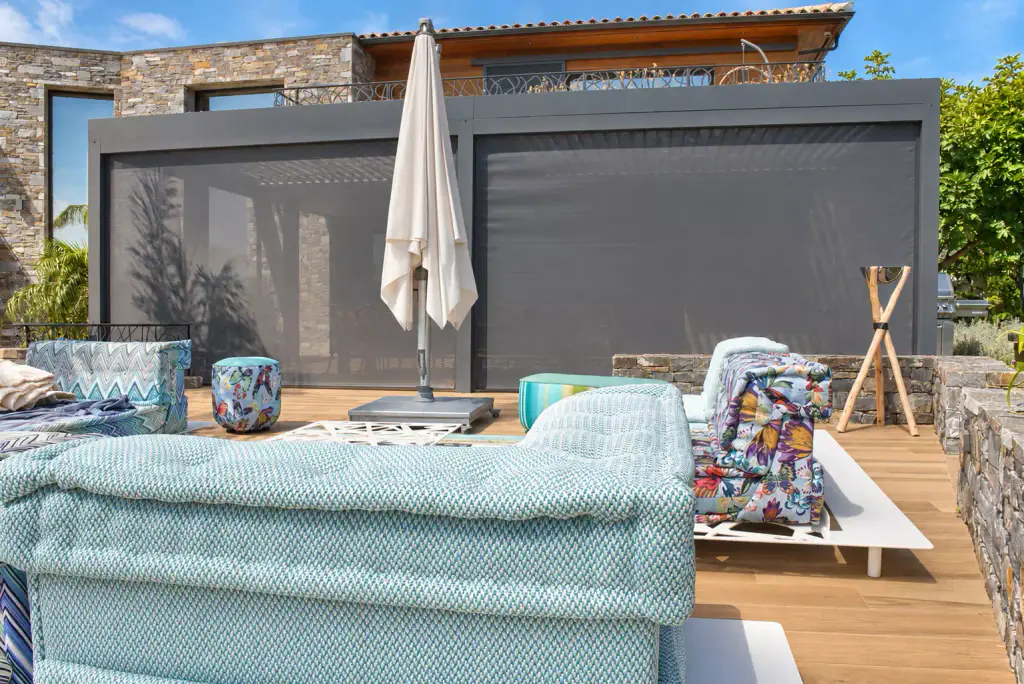 Stores screen gris anthracite entièrement descendus sur deux travées d’une pergola bioclimatique, vus de l’extérieur, salon bas aux tissus turquoise au premier plan.