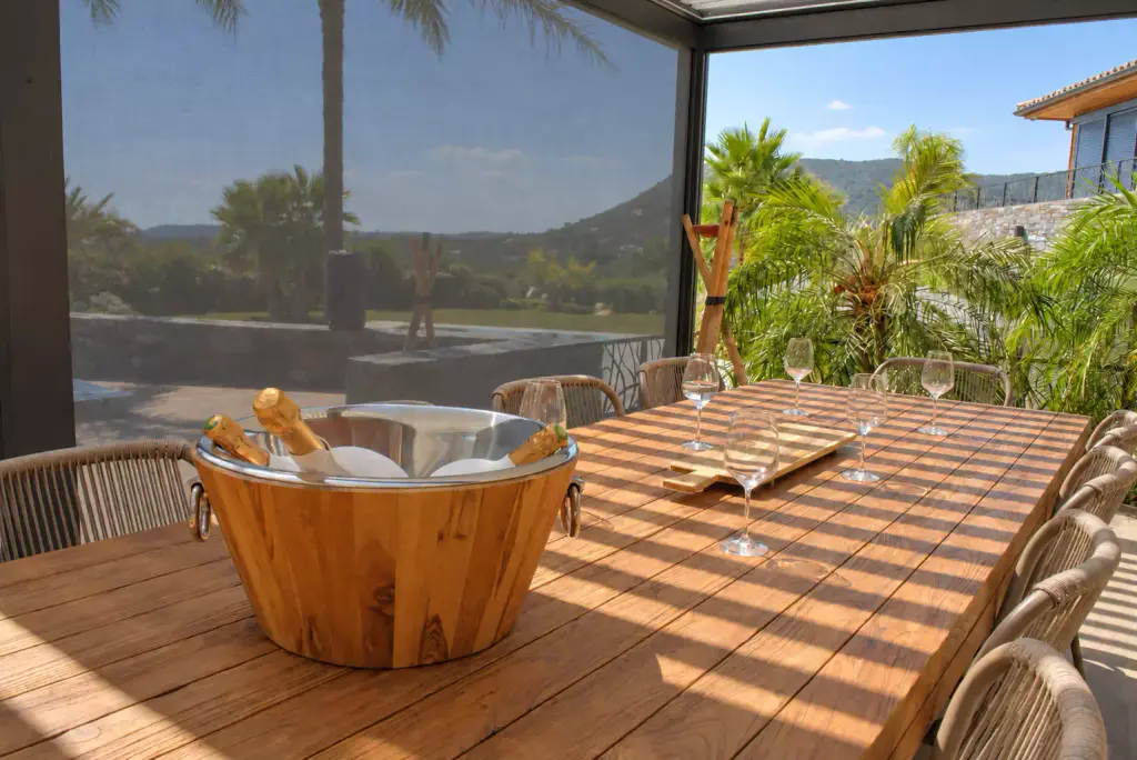 Seau à champagne en bois sur une longue table en bois massif, sous une pergola dont le store screen gris, descendu à gauche, filtre la vue sur les palmiers et les collines.