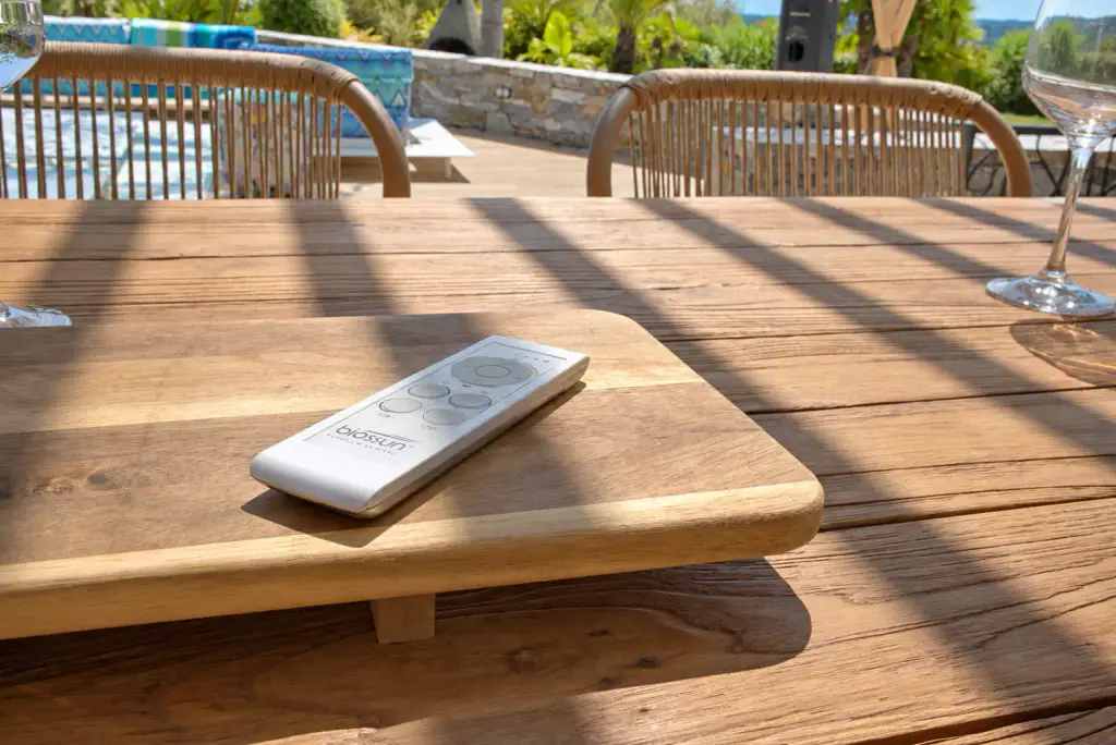 Télécommande blanche de pergola bioclimatique posée sur une planche en bois, sur une table rayée par l’ombre des lames, chaises en corde tressée à l’arrière-plan.