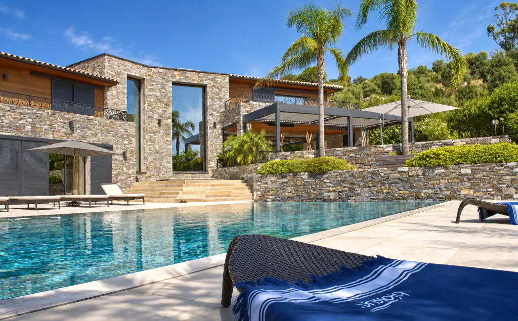 Stone and timber villa beside a turquoise pool, anthracite grey bioclimatic pergola on the upper terrace between two palm trees, sun lounger in the foreground.