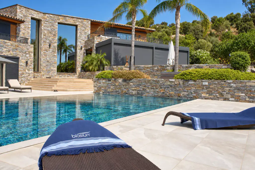Villa en pierre et bois au bord de sa piscine, pergola gris anthracite aux stores screen descendus sur la terrasse haute, serviette bleue sur un bain de soleil au premier plan.