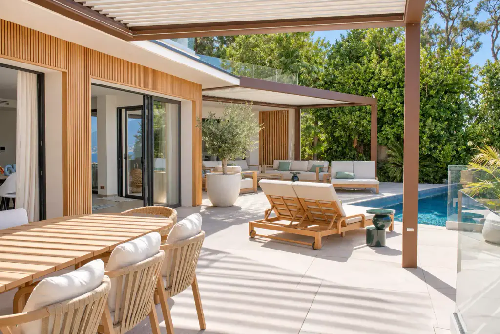 Deux pergolas bioclimatiques brun cuivré le long de la façade en tasseaux de bois : la table en teck sous la première, le salon d’extérieur sous la seconde, la piscine entre.