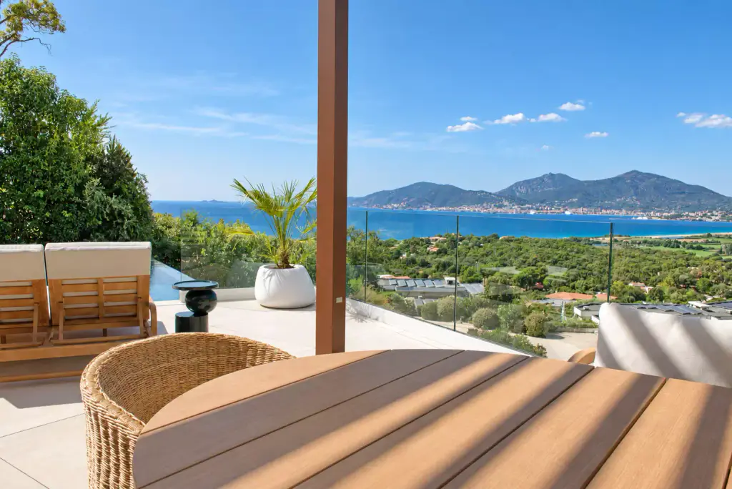 Woven fibre armchair and round teak table in the foreground, copper-brown pergola post; potted palm, glass balustrade, gulf and mountains.