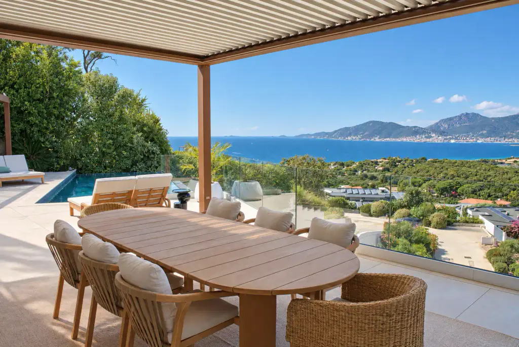 Grande table ovale en teck, chaises en corde et fauteuil tressé sous la pergola bioclimatique aux lames entrouvertes ; piscine à gauche, golfe et montagnes en face.