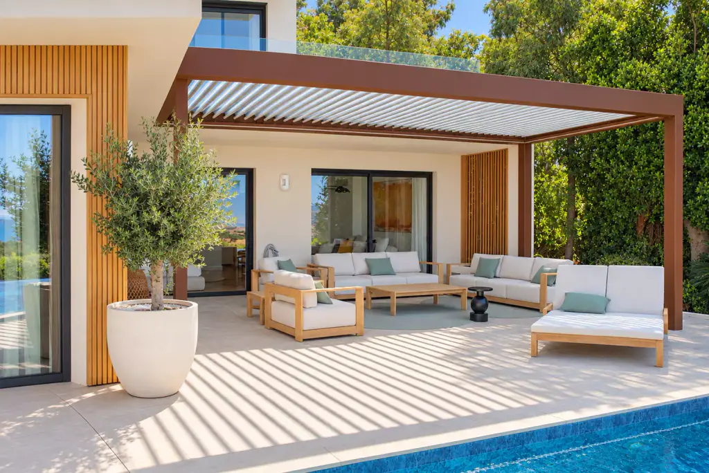 Pergola bioclimatique à structure brun cuivré et lames claires, adossée à la façade : salon d’extérieur en teck et coussins écrus, olivier en pot blanc, bord de piscine.