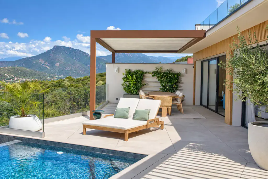 Pergola bioclimatique à cadre brun cuivré et lames fermées, de face : bain de soleil double, table ronde, agrumes palissés au mur et montagne derrière.