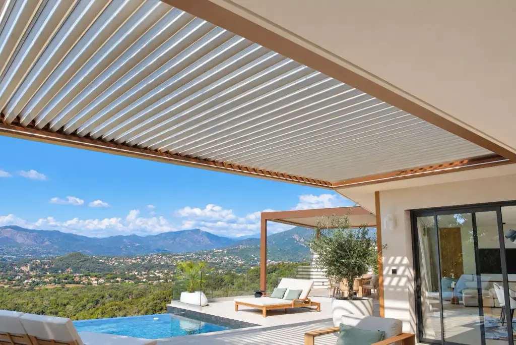 Lames claires et cadre brun cuivré de la pergola en premier plan ; en contrebas du regard, la piscine, les bains de soleil, la seconde pergola et les montagnes.