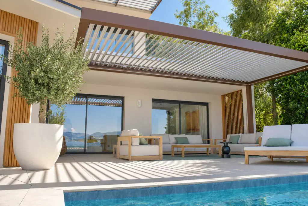 Pergola bioclimatique aux lames claires entrouvertes, vue en contre-plongée depuis la piscine : salon d’extérieur en teck, olivier en pot et casquette à lames à l’étage.