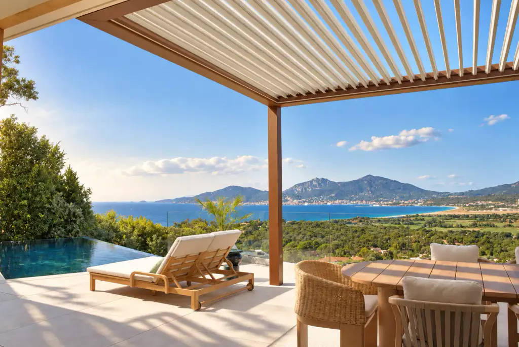 Pergola bioclimatique à structure brun cuivré et lames claires entrouvertes, au-dessus de la table en teck ; bain de soleil, piscine à débordement et golfe au loin.