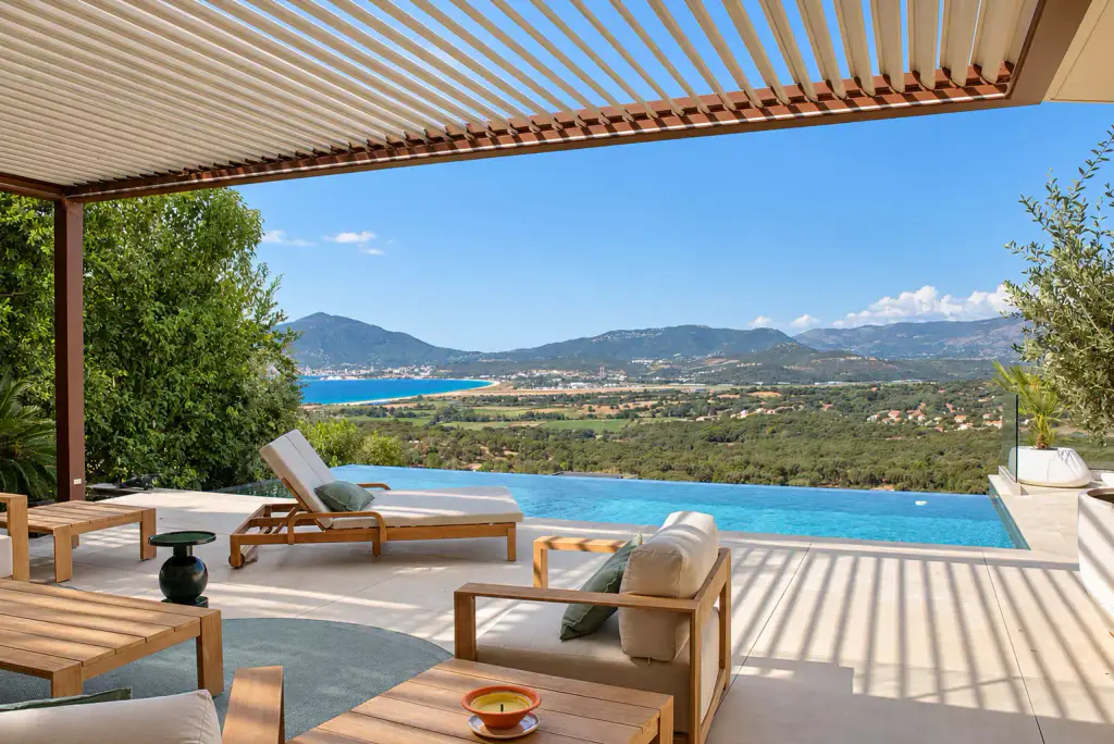 Sous la pergola bioclimatique aux lames claires entrouvertes : fauteuil et tables basses en teck, bain de soleil, piscine à débordement, puis la plaine et le golfe.