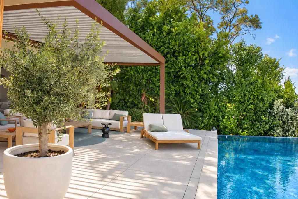 Pergola bioclimatique brun cuivré vue de profil, lames claires inclinées, contre une haie dense : olivier en pot, salon en teck, bain de soleil et angle de piscine.