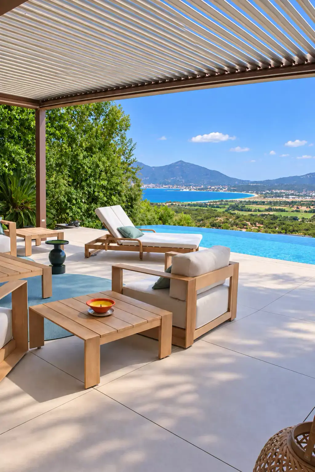Pergola bioclimatique aux lames claires au-dessus du salon d’extérieur : fauteuil et table basse en teck, bain de soleil, piscine à débordement et golfe en face.