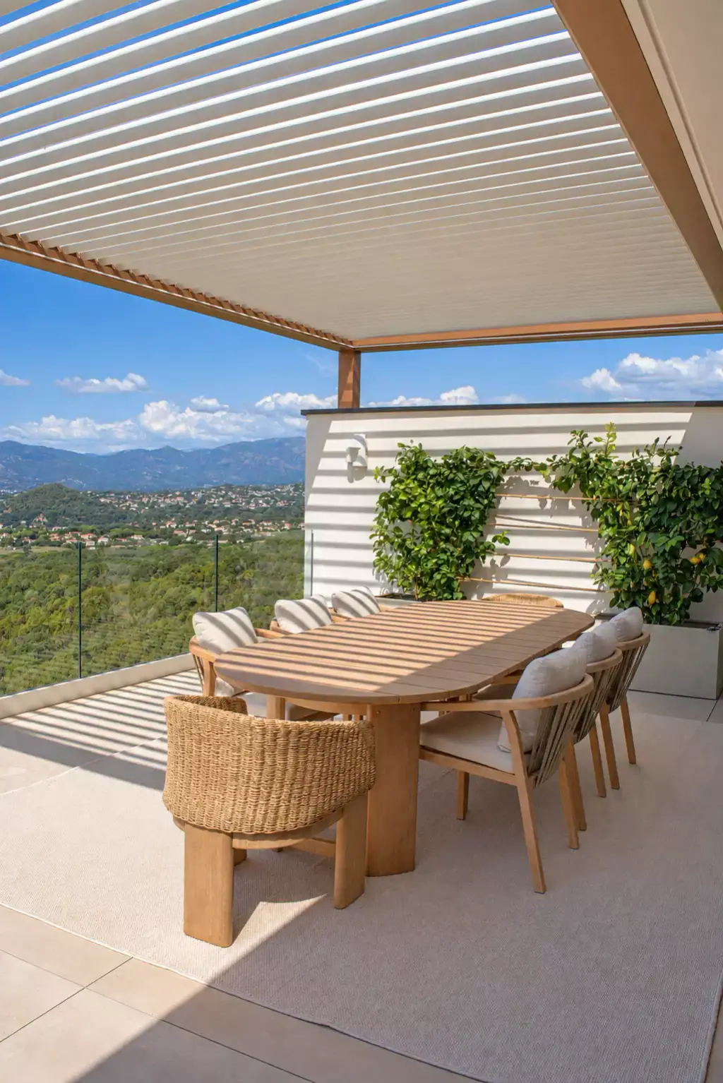 Sous la pergola bioclimatique aux lames claires, table à manger en teck et fauteuil tressé contre le mur aux agrumes palissés ; vallée et montagnes derrière le garde-corps.