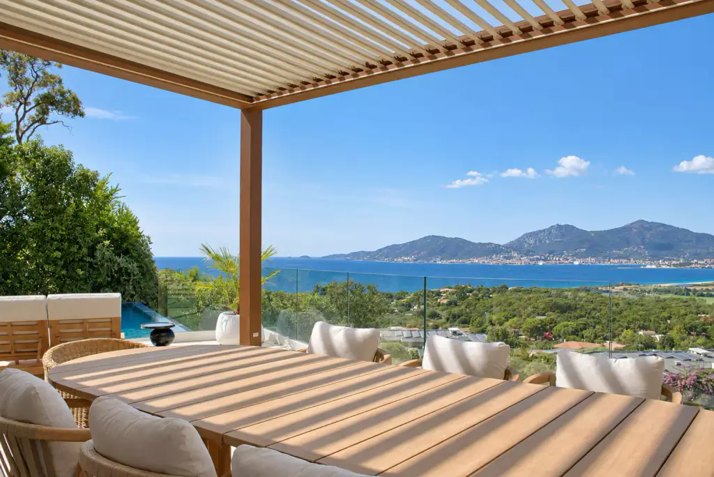 Pergola bioclimatique aux lames claires entrouvertes au-dessus de la grande table en teck ; derrière le garde-corps vitré, le golfe et ses montagnes.