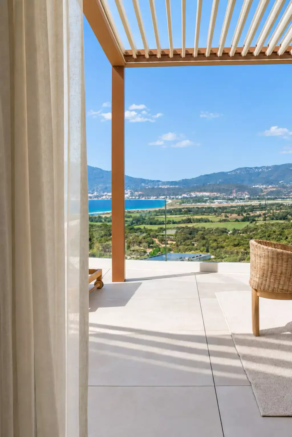 Pergola bioclimatique aux lames entrouvertes vue depuis l’intérieur, un voilage clair à gauche de la baie ; terrasse dallée, fauteuil tressé, plaine et golfe au loin.