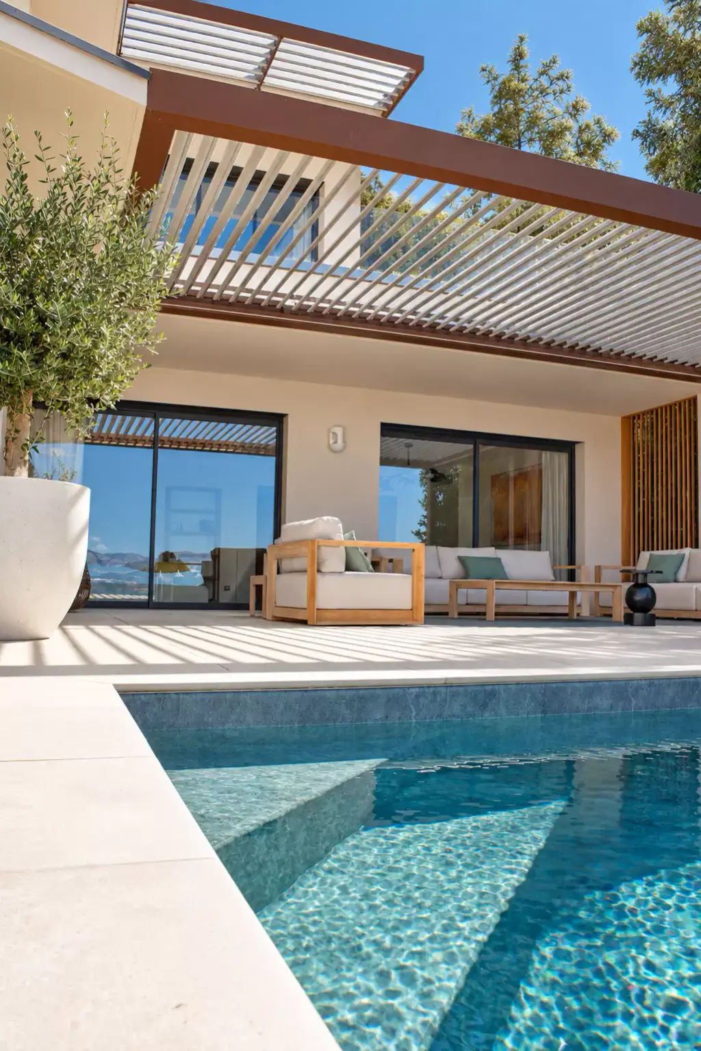 Piscine et sa plage en dalles claires devant la maison : pergola bioclimatique aux lames entrouvertes, salon en teck, olivier en pot et casquette à lames à l’étage.