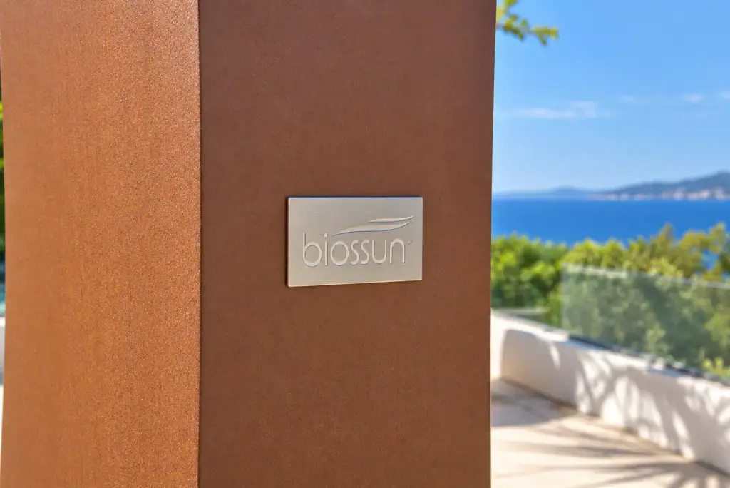 Plaque en métal brossé gravée « biossun » sur un poteau de pergola au thermolaquage brun cuivré texturé ; la mer et le garde-corps vitré, flous, à droite.