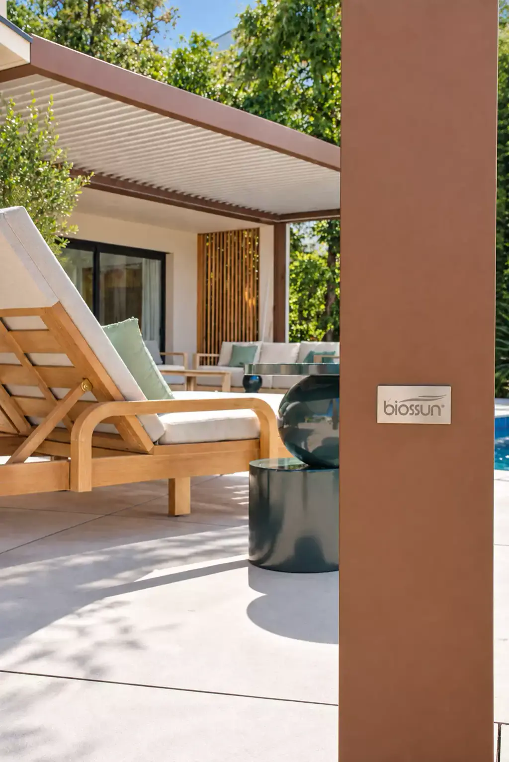 Poteau de pergola brun cuivré portant la plaque en métal brossé du fabricant Biossun ; derrière, flous, un bain de soleil en teck et le salon d’extérieur.