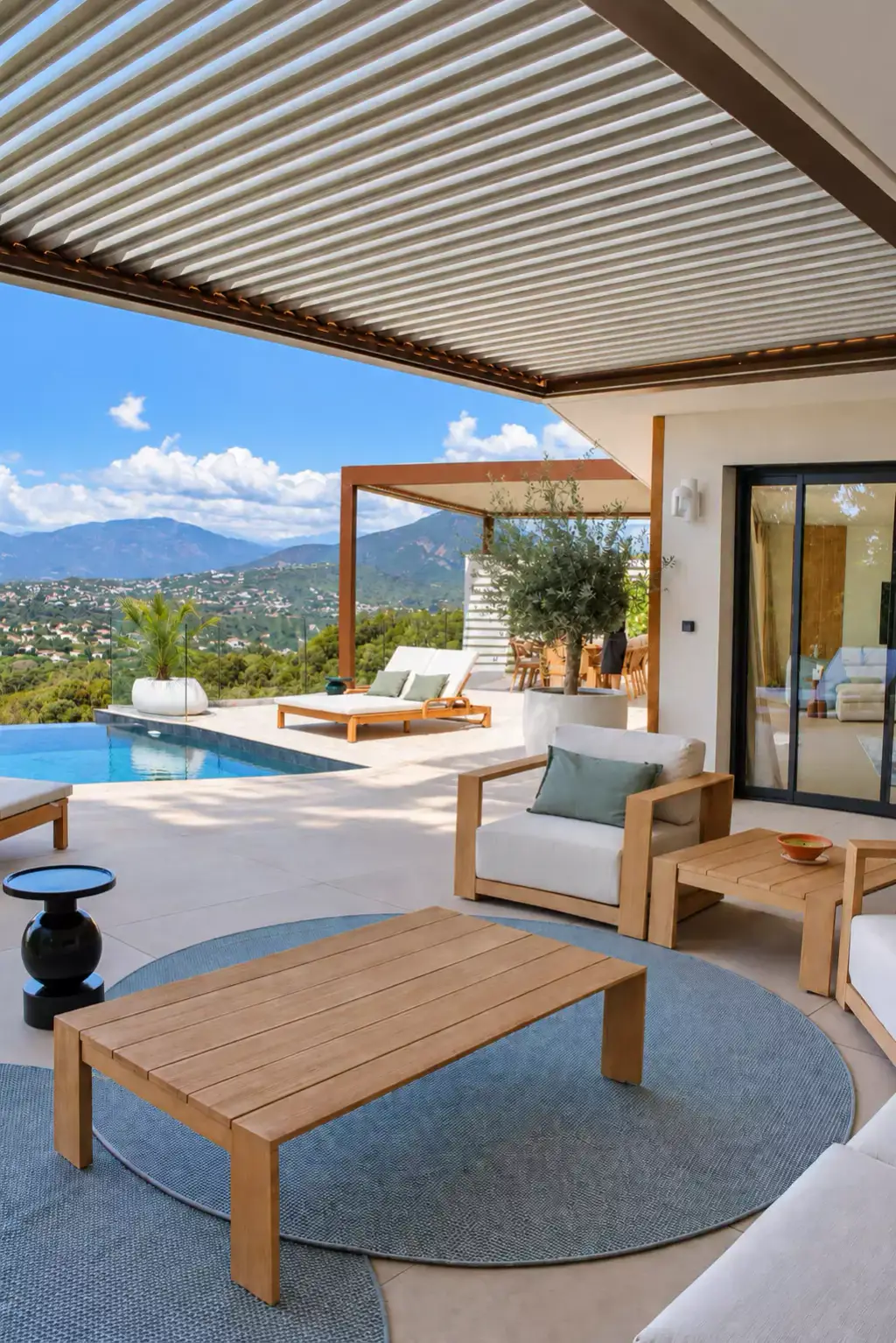 Salon d’extérieur sous la pergola : table basse en teck sur un tapis rond bleu-vert, fauteuil aux coussins écrus ; au fond, la piscine et la seconde pergola.